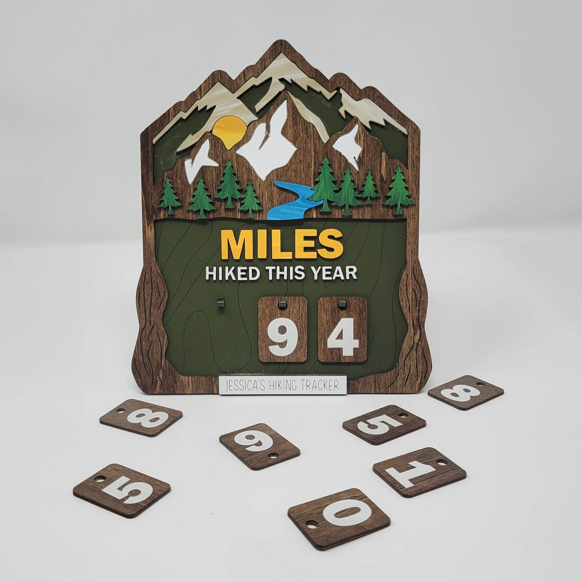 Layered wooden hiking tracker sign featuring mountains, evergreen trees, a river, and sunrise, with the text “Miles Hiked This Year” and interchangeable number tiles