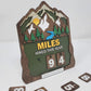 Layered wooden hiking tracker sign featuring mountains, evergreen trees, a river, and sunrise, with the text “Miles Hiked This Year” and interchangeable number tiles at an angle