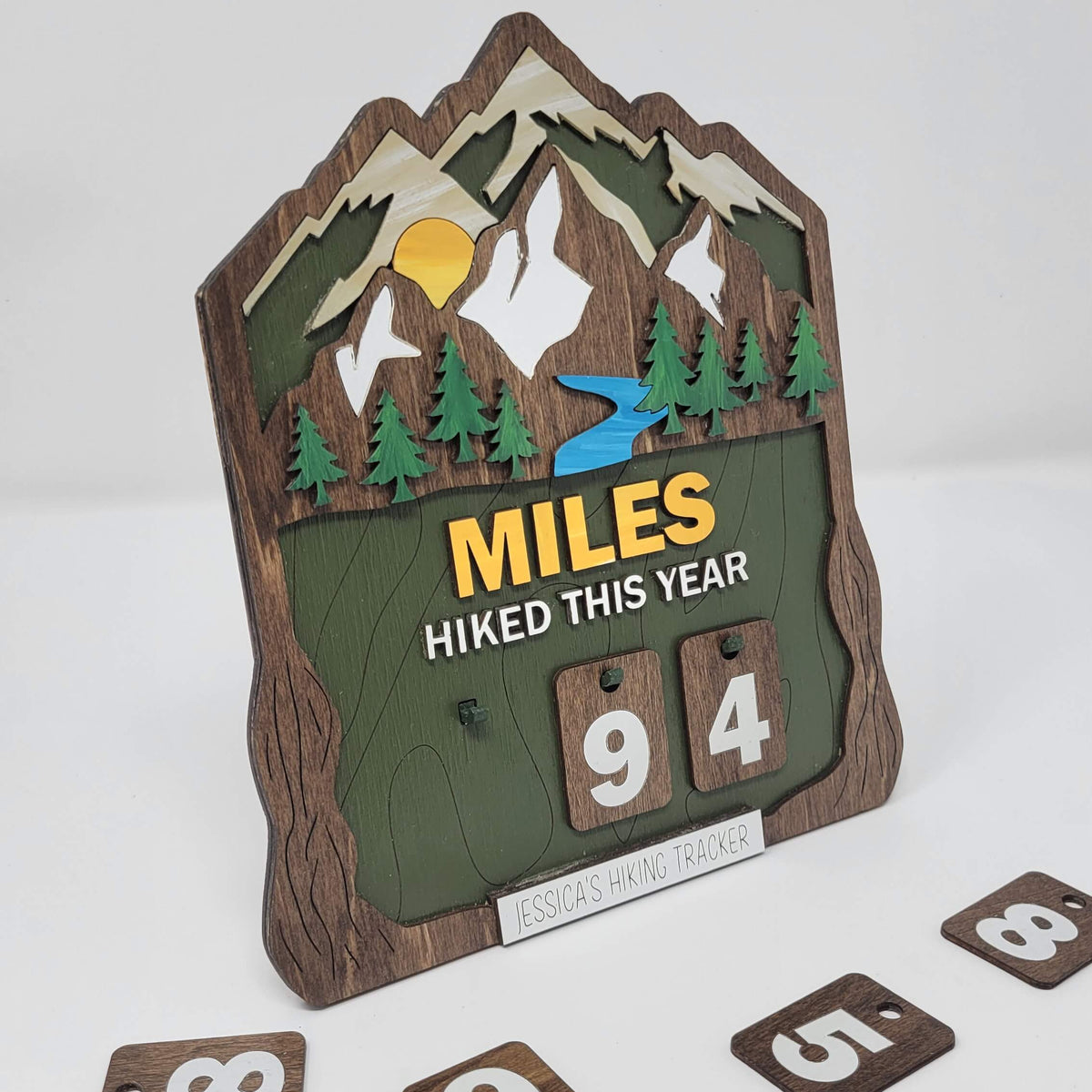 Layered wooden hiking tracker sign featuring mountains, evergreen trees, a river, and sunrise, with the text “Miles Hiked This Year” and interchangeable number tiles at an angle